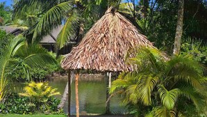 Une construction à toit de chaume, entourée d’une végétation luxuriante et bordée par un plan d’eau.