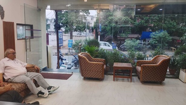 A man sitting in a chair in a room with a view of the street outside. There are two patterned armchairs and a small table in the room. - Hotel Swaagat (Agra)