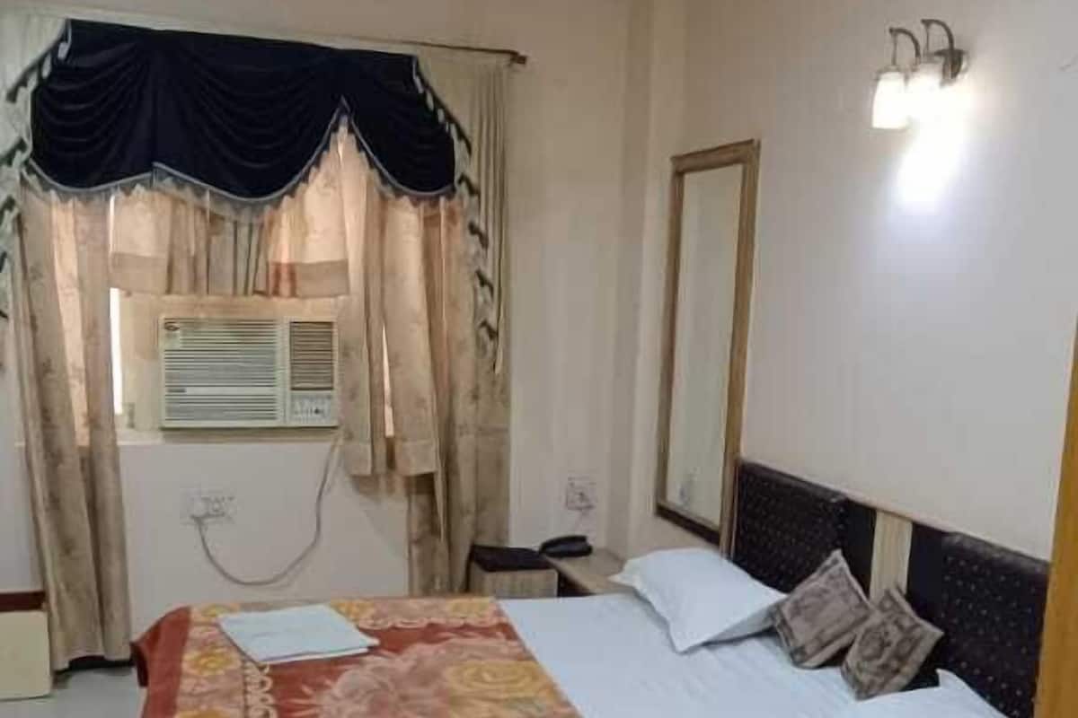 A hotel room with a bed, a ceiling fan, a window with curtains, a wall-mounted light, and a bedside table.