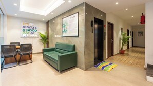 A modern reception area with a green sofa, black chairs, a reception desk, a framed informational poster, and a potted plant. - Treebo Flora Embassy (Chennai)