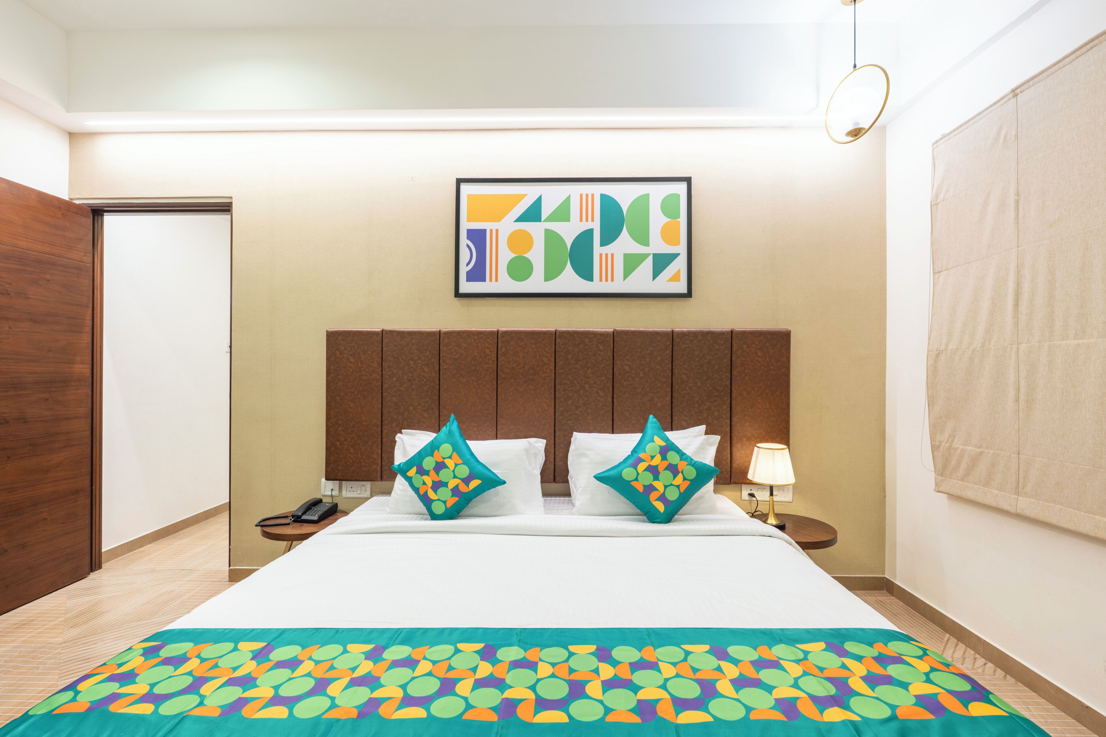 Quarto de hotel com uma cama, duas almofadas, mesa de cabeceira com abajur, telefone e uma obra de arte na parede.
