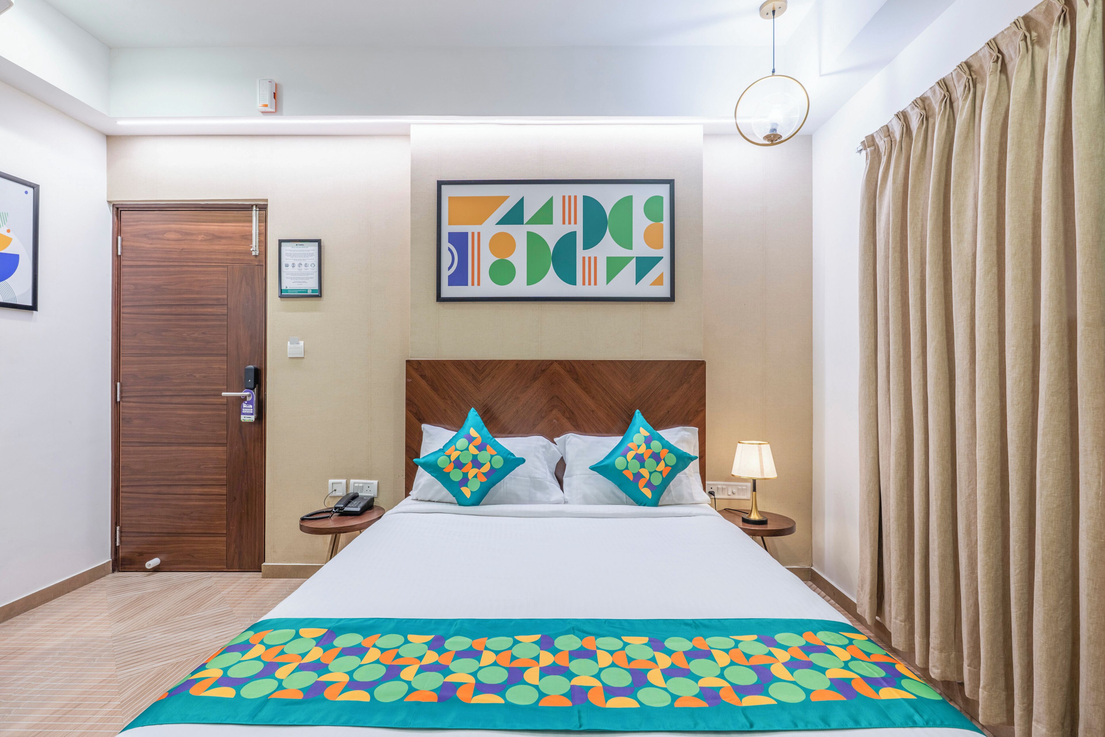 A hotel room with a bed, a wooden headboard, a framed abstract artwork, a bedside table with a lamp, and a door.