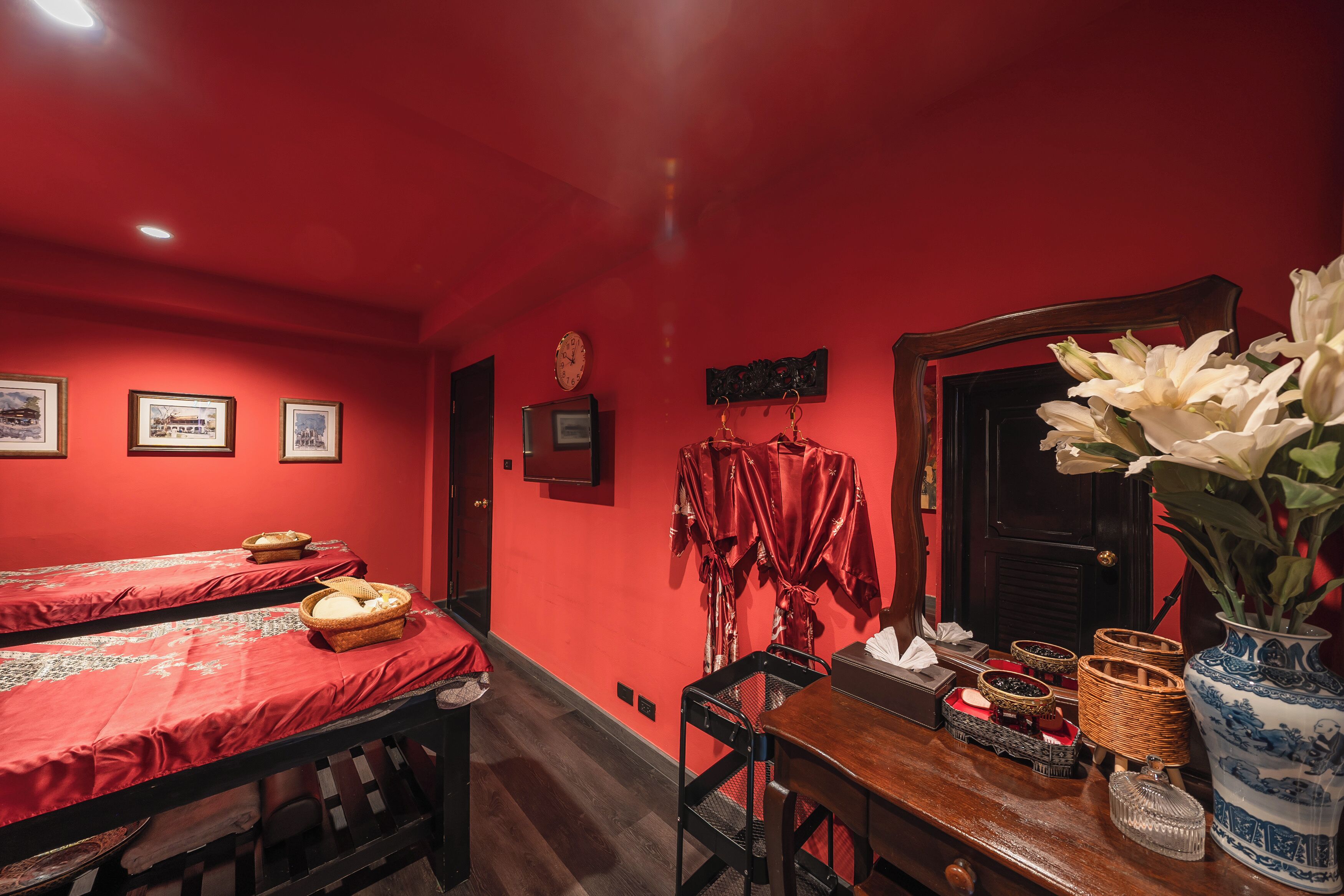 Une pièce aux murs rouges, avec deux lits recouverts de draps rouges, une table en bois sur laquelle sont posés des paniers, un vase rempli de fleurs et une horloge accrochée au mur.