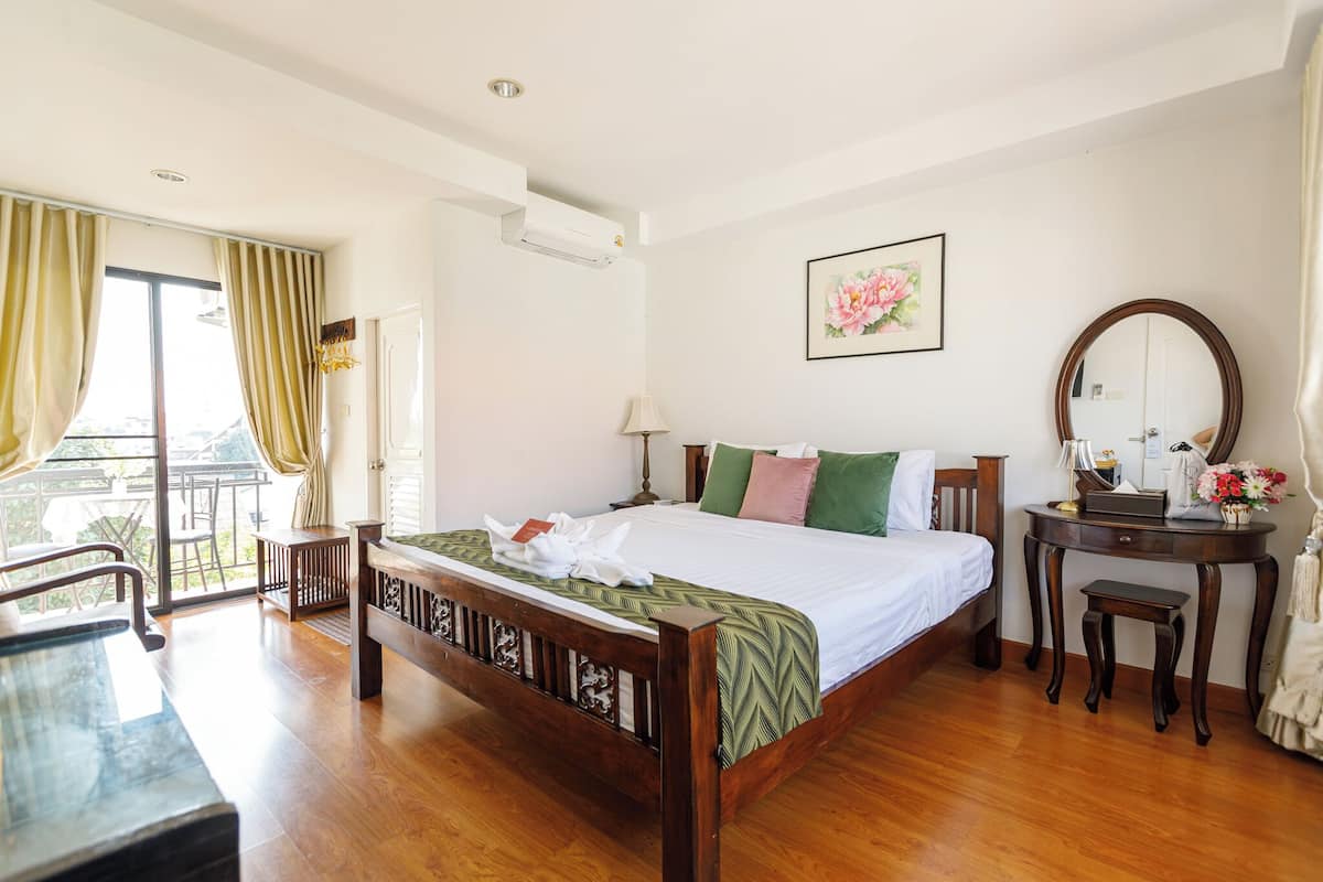 Habitación de hotel con cama de madera, un escritorio, una silla, un balcón con cortinas, aire acondicionado de pared, un cuadro floral enmarcado y un espejo redondo sobre una mesita de noche.