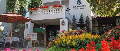 A building with a sign reading "Rachamankha Flora House" surrounded by vibrant flower beds.