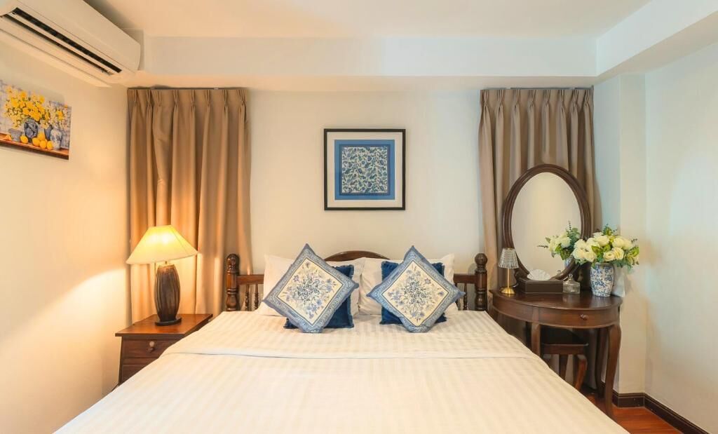 A neatly arranged hotel room with a bed, a bedside table with a lamp, a mirror, a framed artwork on the wall, and a vase with flowers.