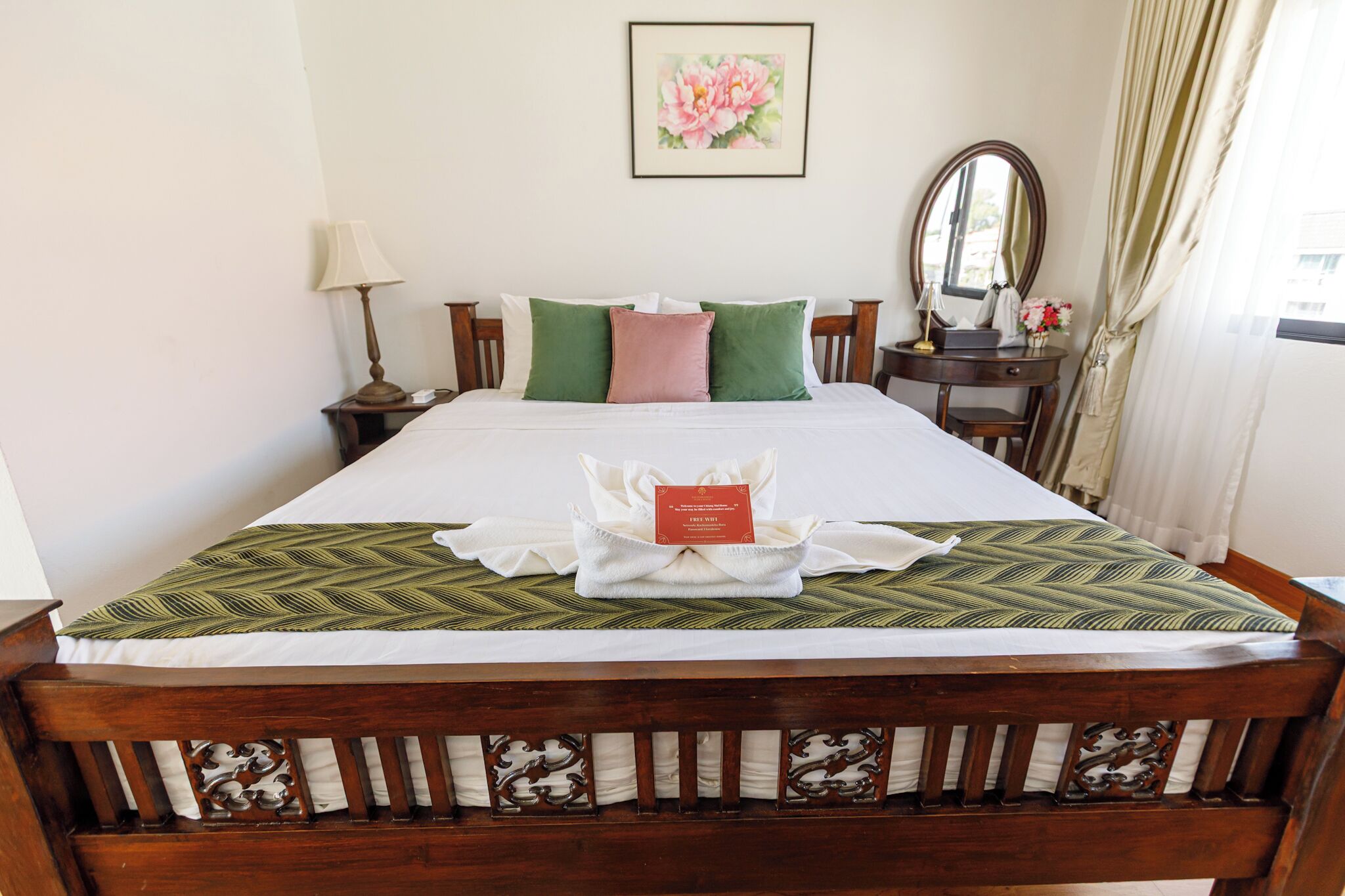 A neatly made bed with a green and white bedspread, a wooden headboard with intricate carvings, a bedside table with a lamp, and a framed floral artwork on the wall.