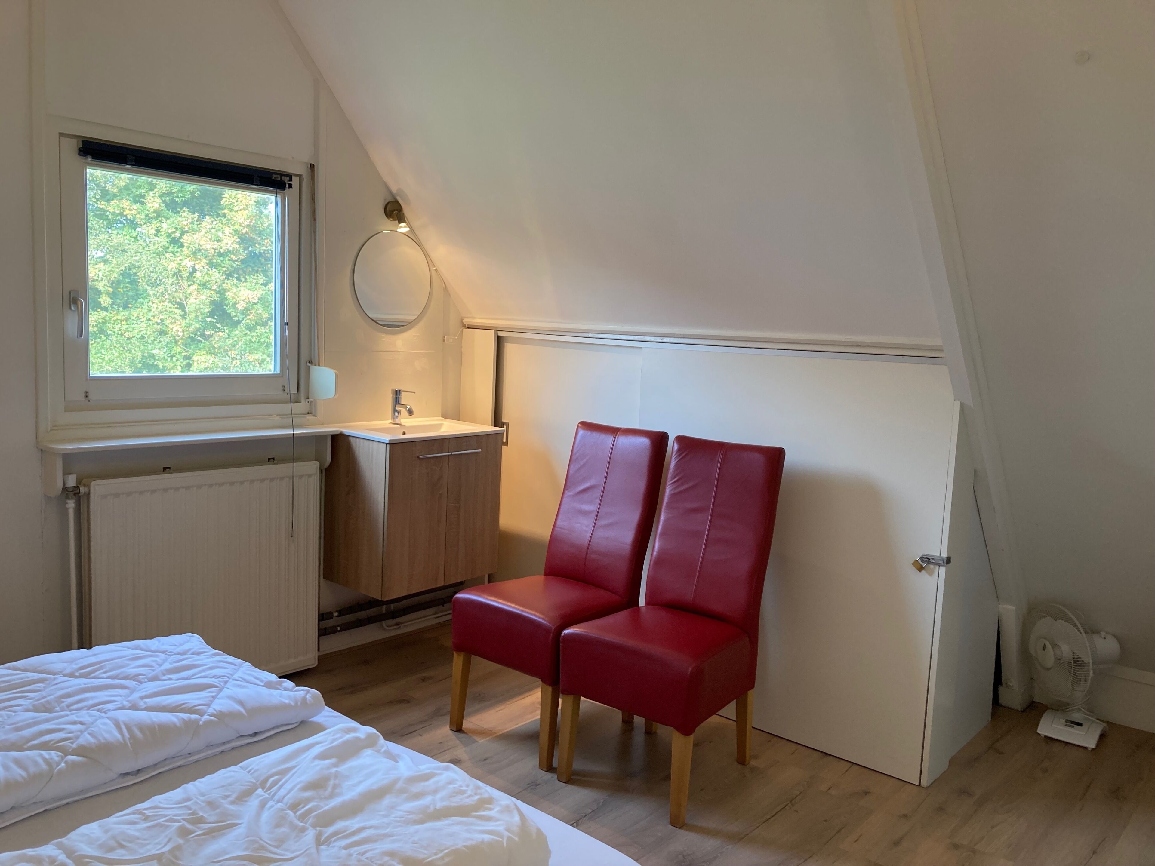 Een slaapkamer met een bed, een rode stoel, een kleine badkamer met een wastafel en een raam.