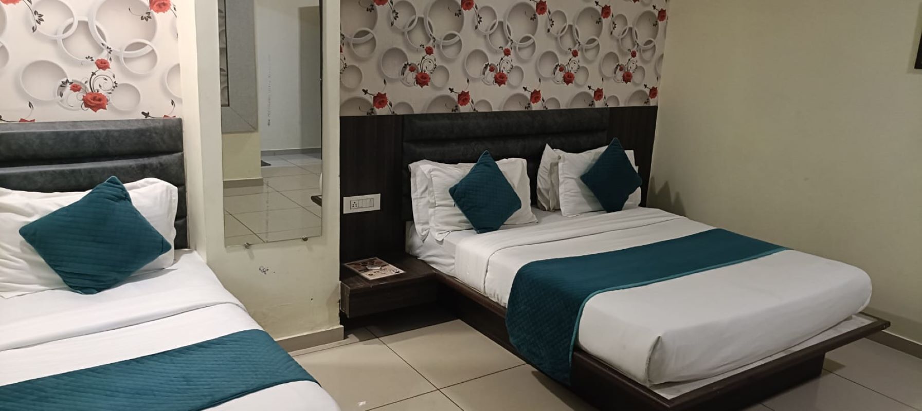 Habitación de hotel con dos camas, cada una con ropa blanca y almohadas color turquesa. La habitación presenta un diseño moderno con papel tapiz estampado y un cabecero oscuro.