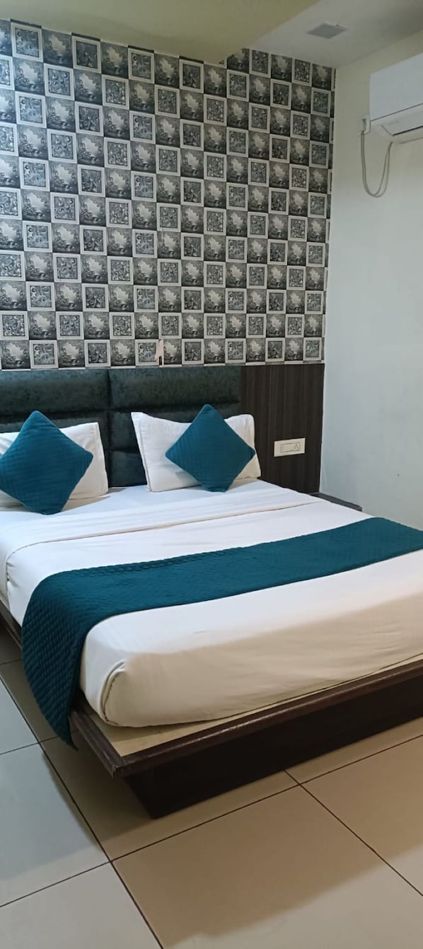A neatly made bed with a teal throw blanket and pillows, a patterned wallpaper headboard, and a white air conditioning unit on the wall. - HOTEL SHIV RESIDENCY (Vadodara)