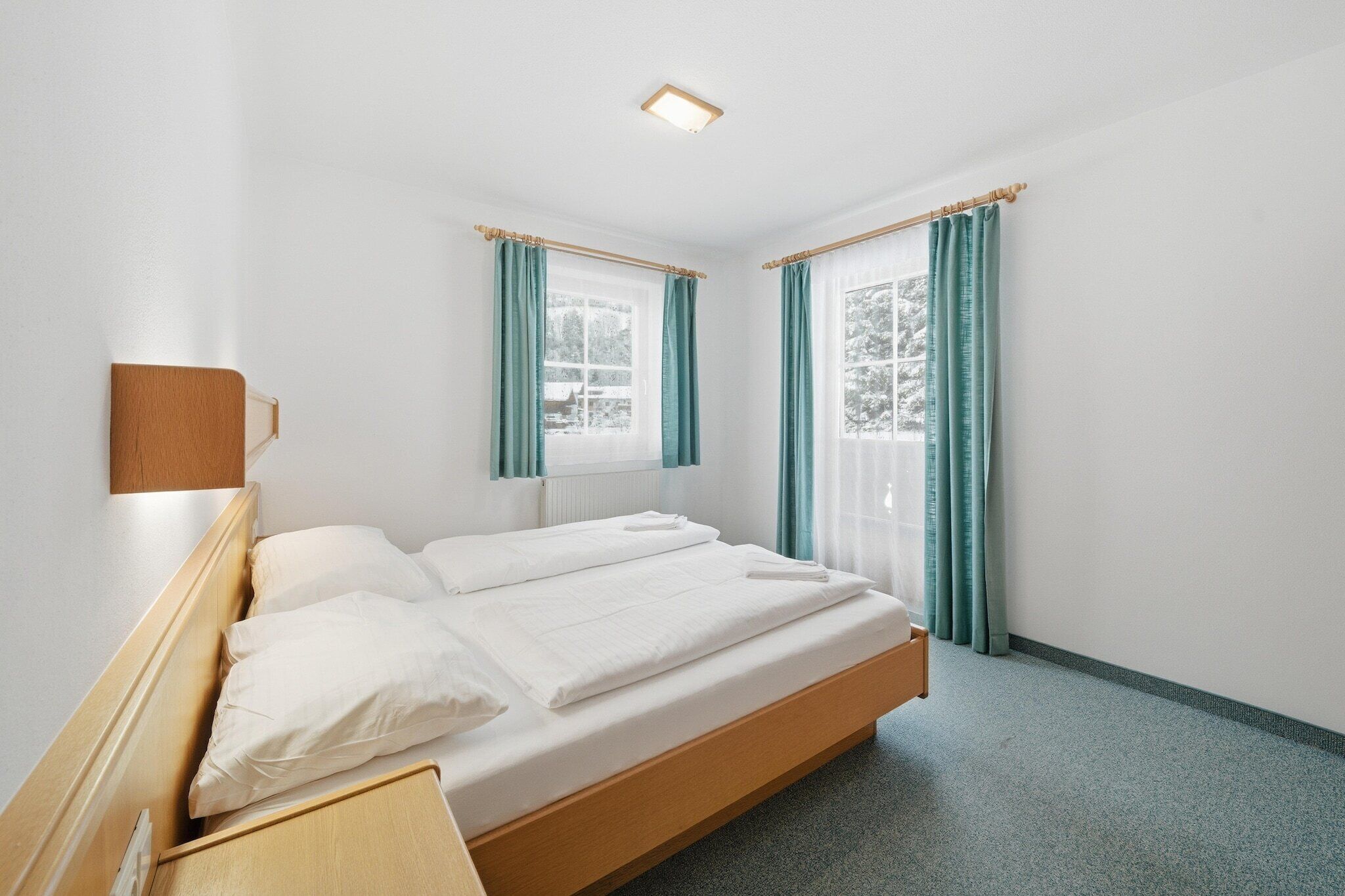 Une chambre d’hôtel propre et moderne, avec un lit simple, deux fenêtres avec rideaux et une petite table de chevet.