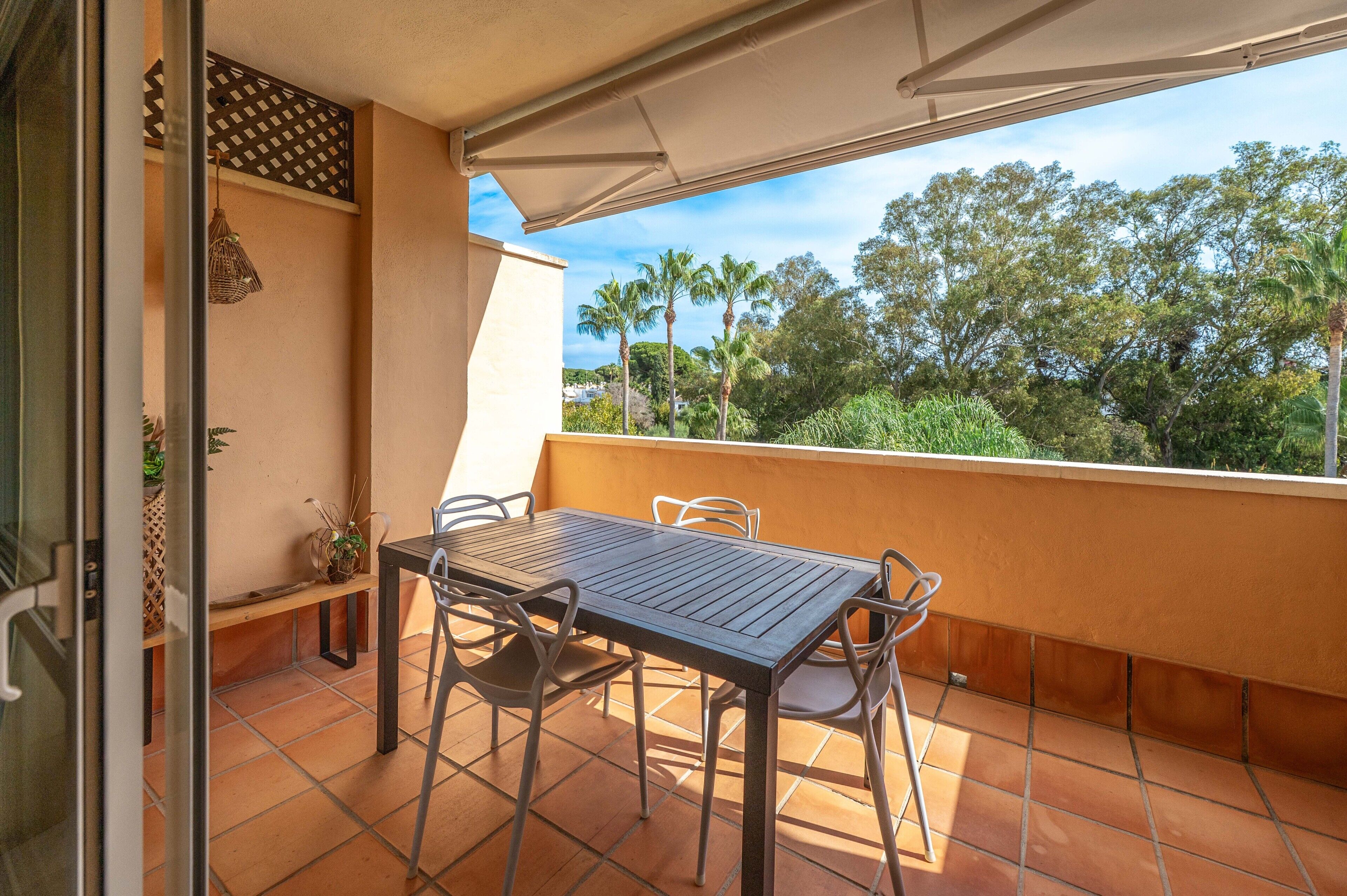 A balcony with a table and chairs, a hanging light fixture, and a view of trees and sky.