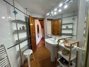 Une salle de bain avec des murs recouverts de carreaux blancs, un lavabo blanc, des étagères, une porte en bois et un miroir.