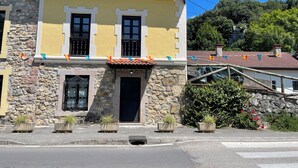 A two-story building with a stone facade, a yellow upper section, and a tiled roof. There are potted plants in front of the entrance, and colorful flags are strung across the top. - La Casina del Arroyo, rural cottage for 2-4 people in Poo de Cabrales (Póo de Cabrales)