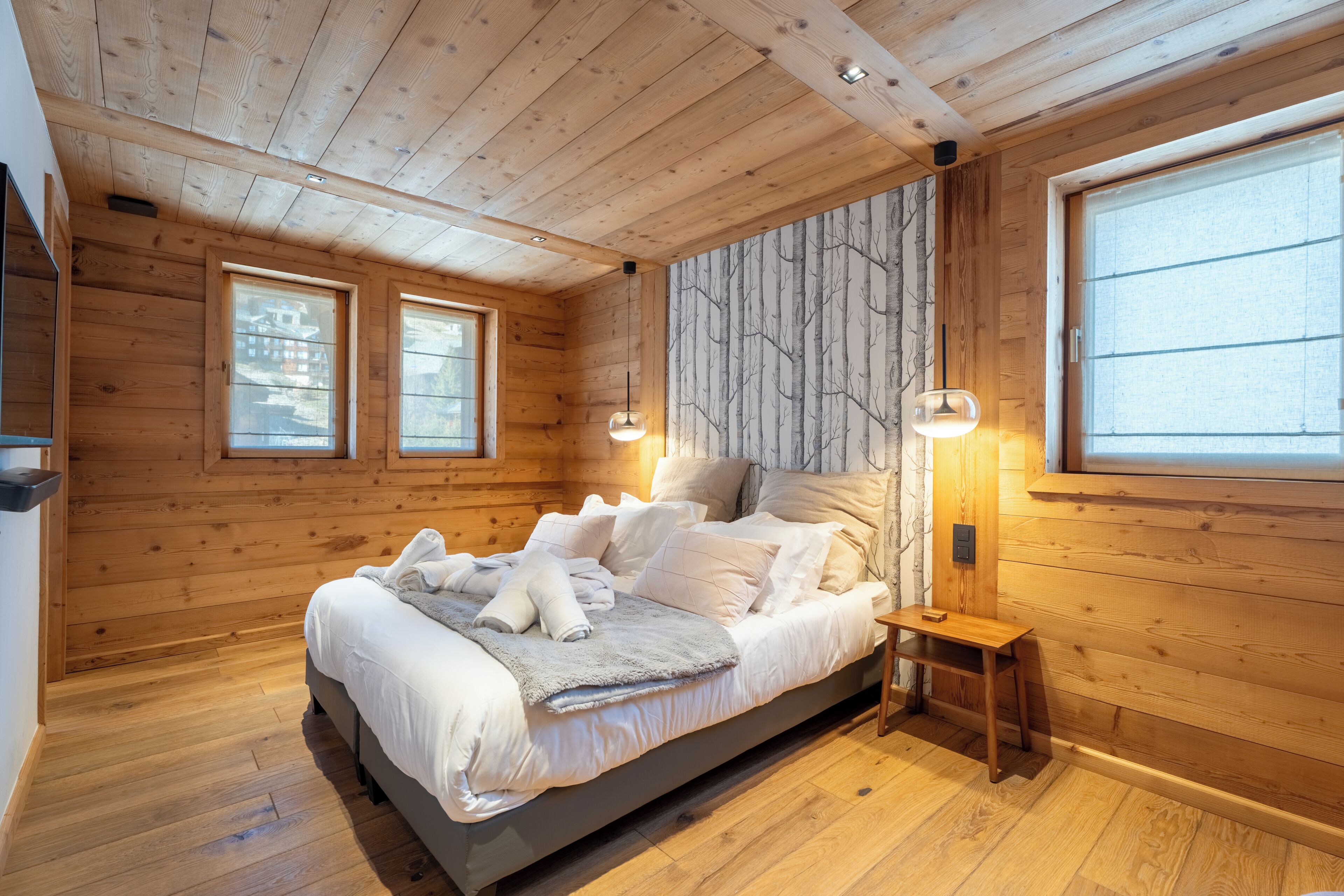 Une chambre dans une cabane en bois, avec un lit, deux fenêtres, une table de chevet et une fresque murale.