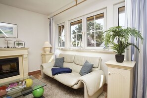 A living room with a sofa, a fireplace, a window with curtains, and a potted plant. - Spacious vacation apartment with terrace and garden in the quiet Landhaus Wildrose - (Zirkow)