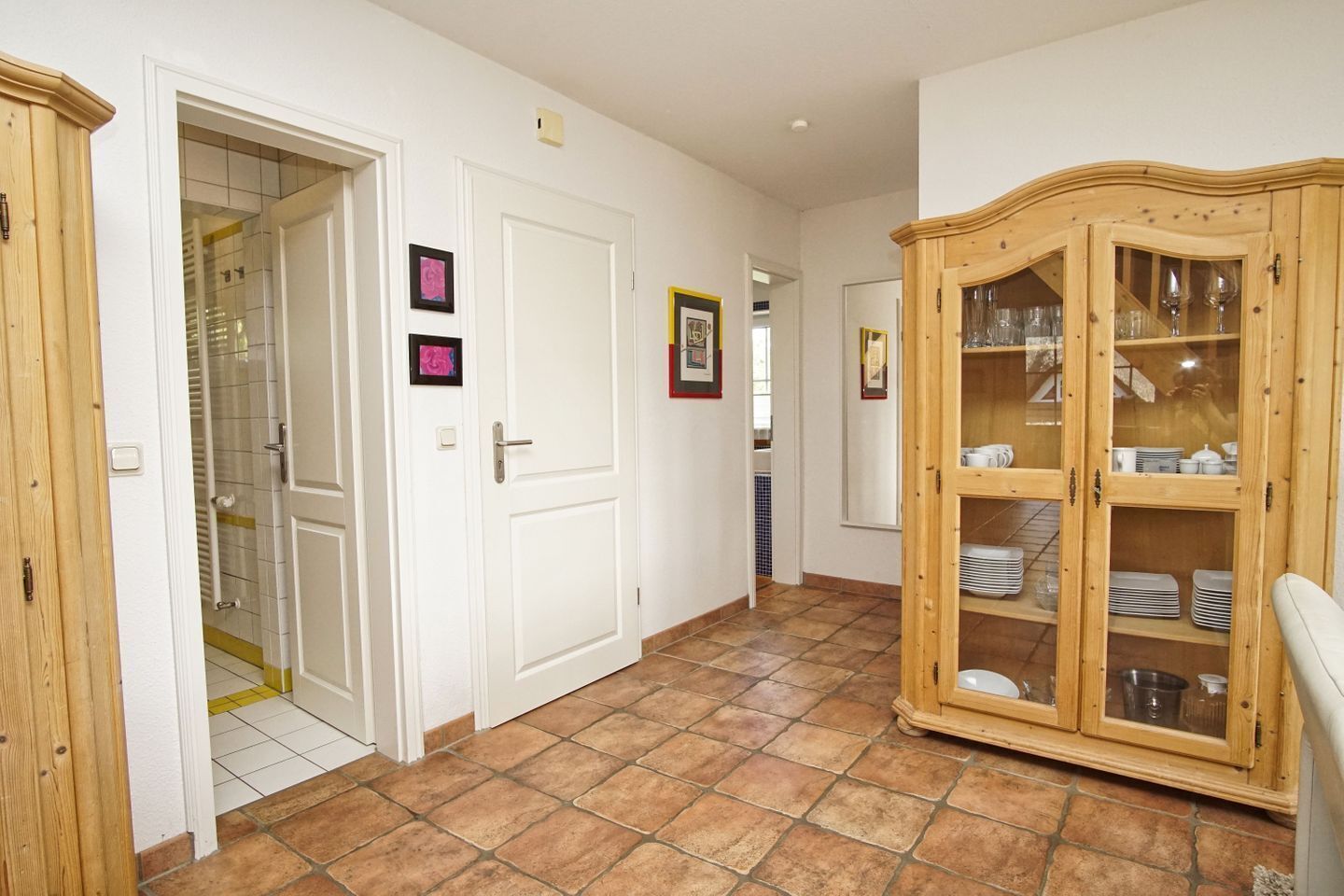 A hallway with a wooden cabinet containing plates and glasses, a white door, and a tiled floor.