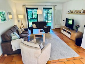 A living room with a sofa, armchair, coffee table, TV, and a window with curtains. - Ferienhaus Boddenblick - Charming semi-detached house in Trips on Rügen - (Bergen auf Rügen)