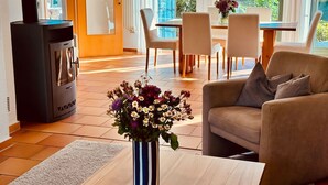 A modern living room with a sofa, a wooden coffee table, a striped vase with flowers, a black wood-burning stove, a dining area with chairs and a table, and large windows with a view of greenery.
