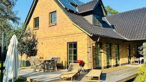A two-story house with a sloped roof, solar panels, a wooden deck with outdoor furniture, and a brick exterior.