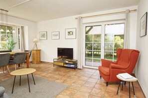 A living room with a TV, a red armchair, a dining table with chairs, and a view through a glass door to a garden.