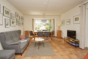A living room with a grey sofa, a dining table with chairs, a TV, and framed pictures on the walls.