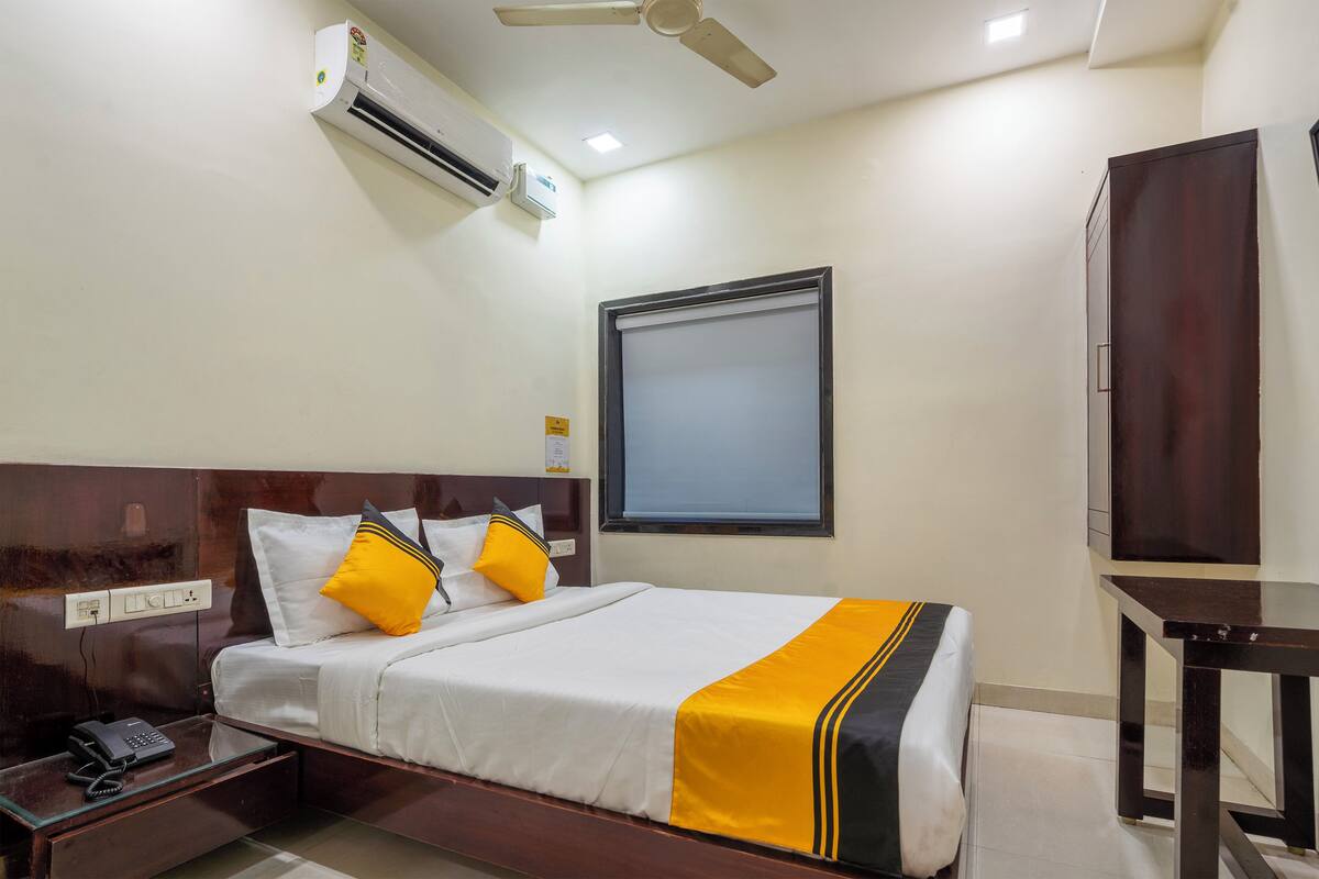 A hotel room with a bed, bedside table, phone, air conditioning unit, ceiling fan, window, and wardrobe.