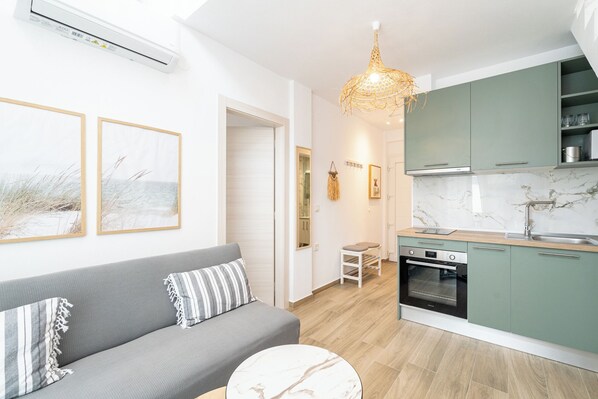 Un appartement moderne avec un canapé gris, une petite table à manger, une cuisine aux meubles verts et un luminaire suspendu.