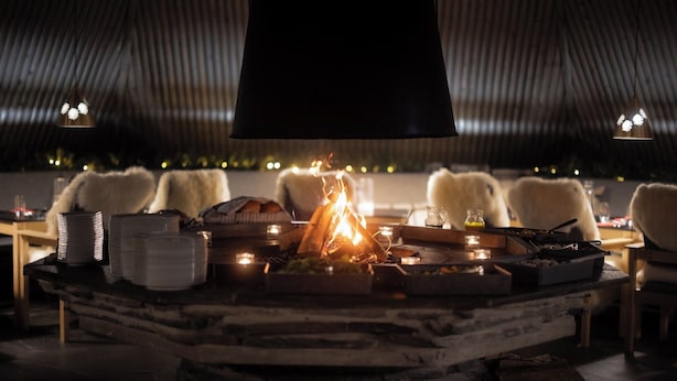 Un coin repas chaleureux avec un foyer central, entouré de chaises blanches et d’une table dressée avec des assiettes et des bougies.