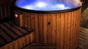 Un bain à remous en bois, accessible par des marches, entouré d’une clôture en bois et d’arbres.