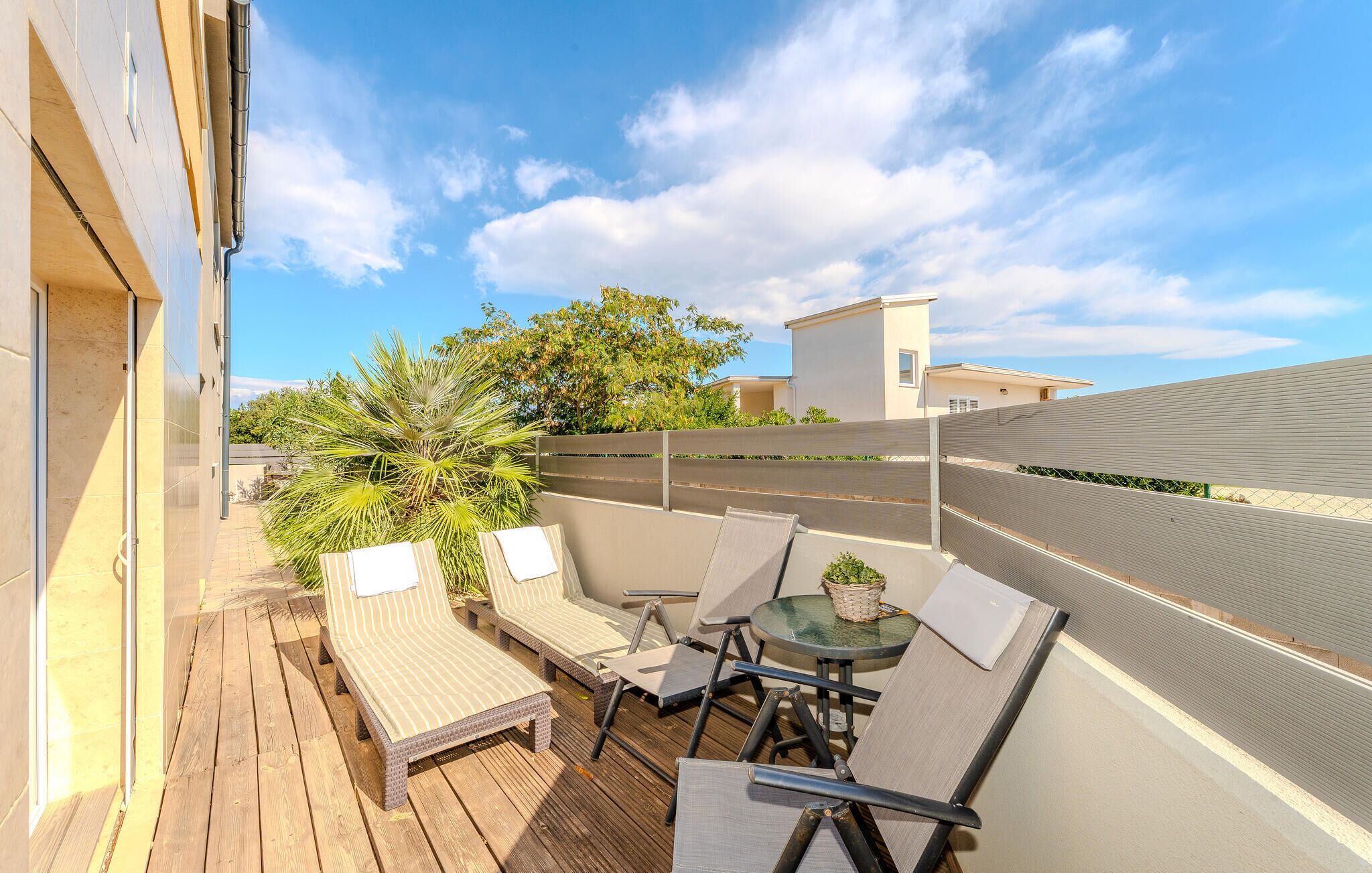Eine Holzterrasse mit Gartenmöbeln, darunter Liegestühle und ein Tisch, mit Blick auf ein Wohngebiet und einen klaren Himmel.