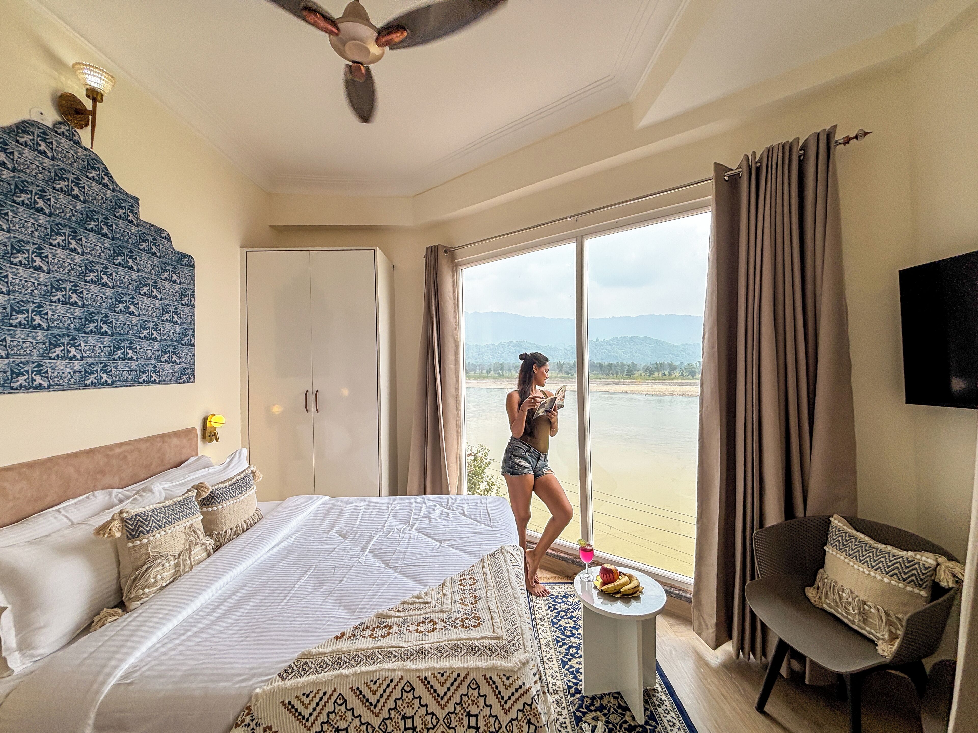 A hotel room with a bed, a small table with a drink, a chair, and a view of a river.