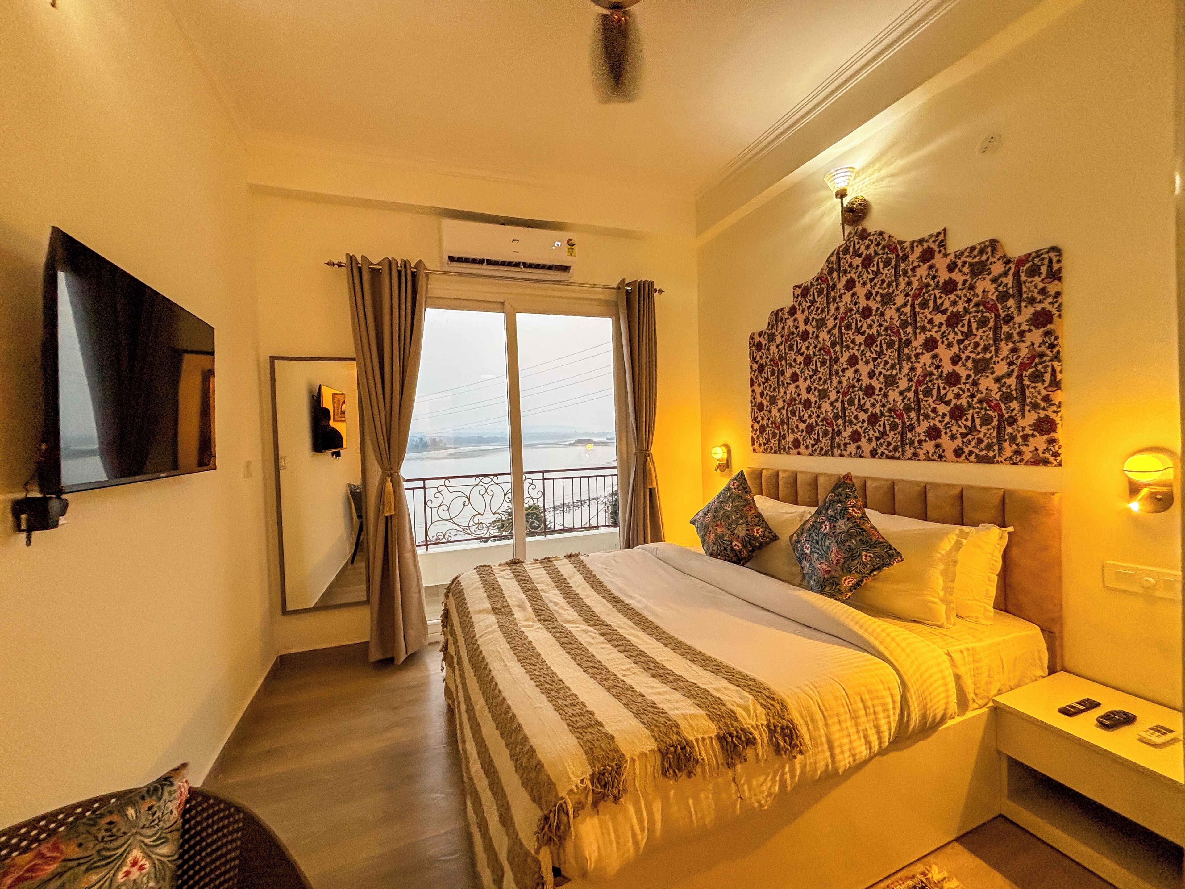 A hotel room with a large bed, a flat-screen TV, a balcony with curtains, and a decorative wall piece.