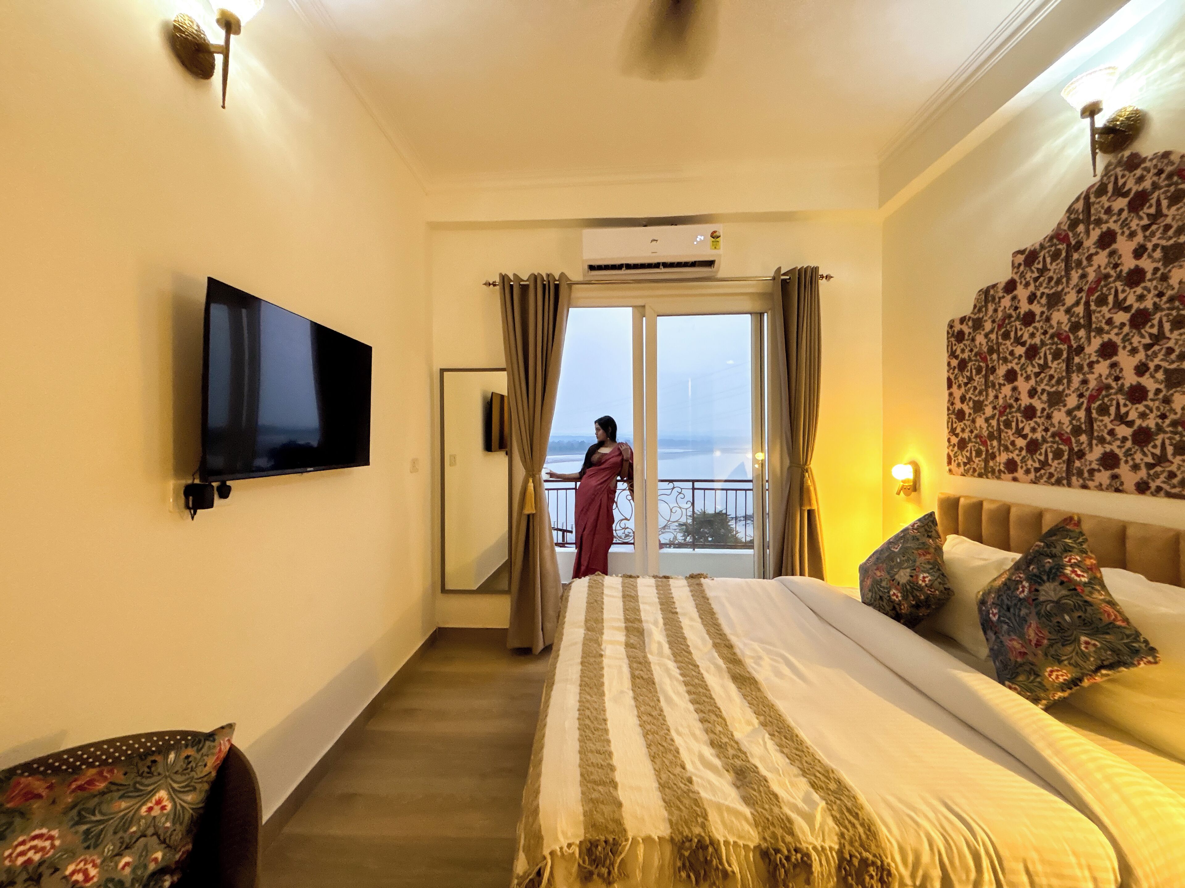 A hotel room with a bed, a flat-screen TV, a balcony with a view, and a ceiling fan.