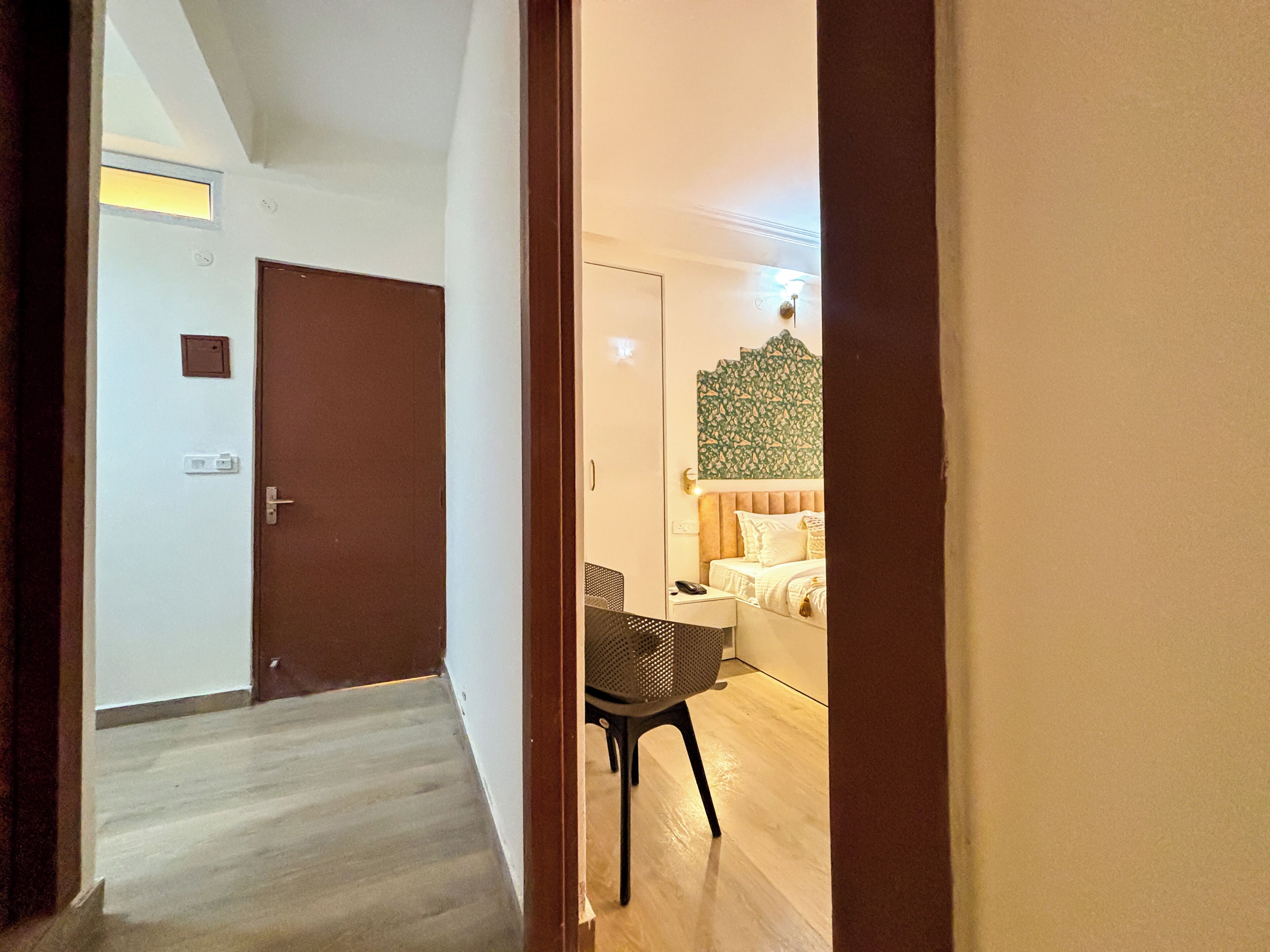 Habitación de hotel con piso de madera, cama con ropa blanca, un escritorio pequeño con silla y una lámpara fijada a la pared.