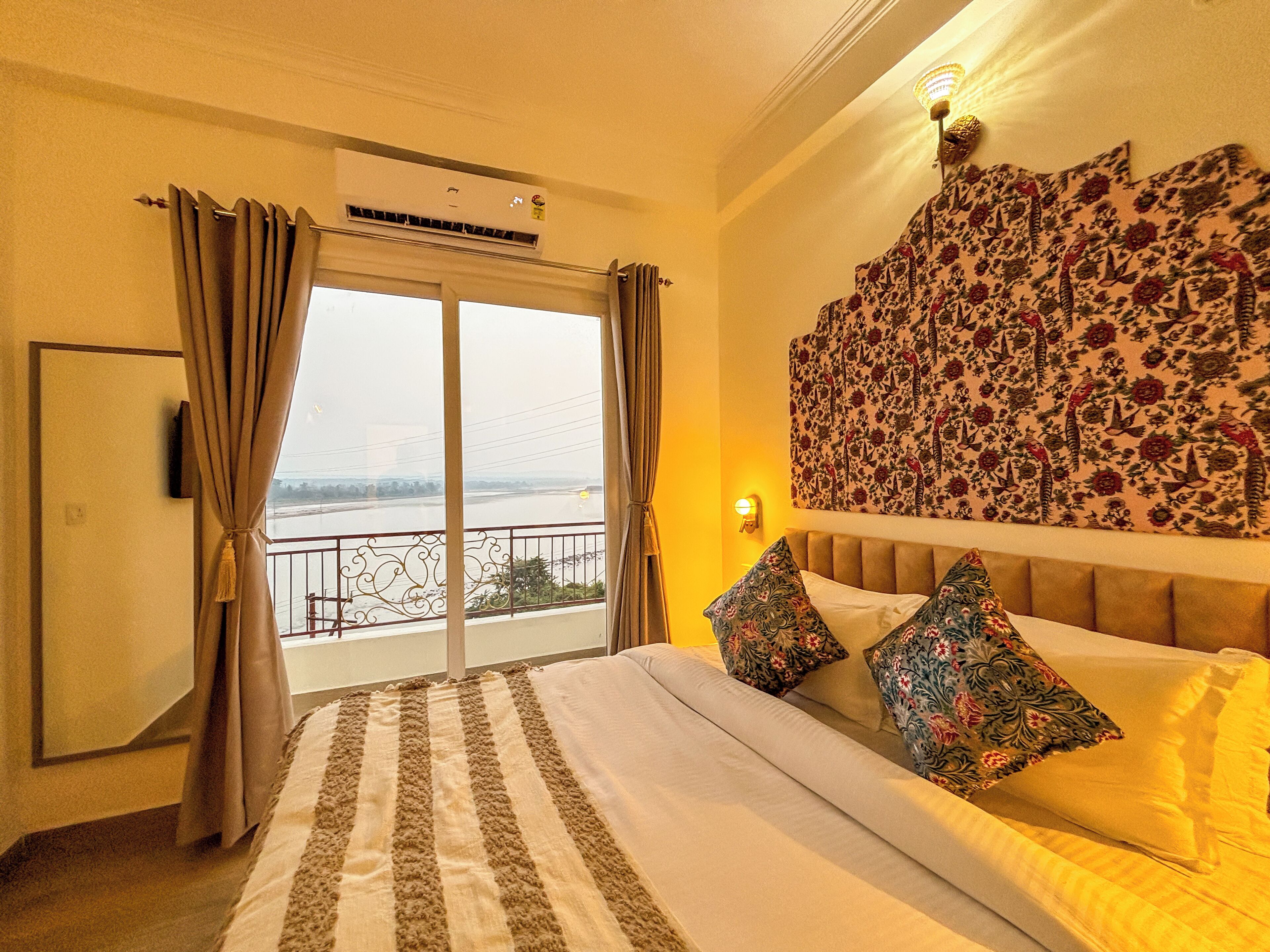 Una habitación de hotel con cama, cortinas, aire acondicionado, vista al río y cabecera tapizada con motivos florales.