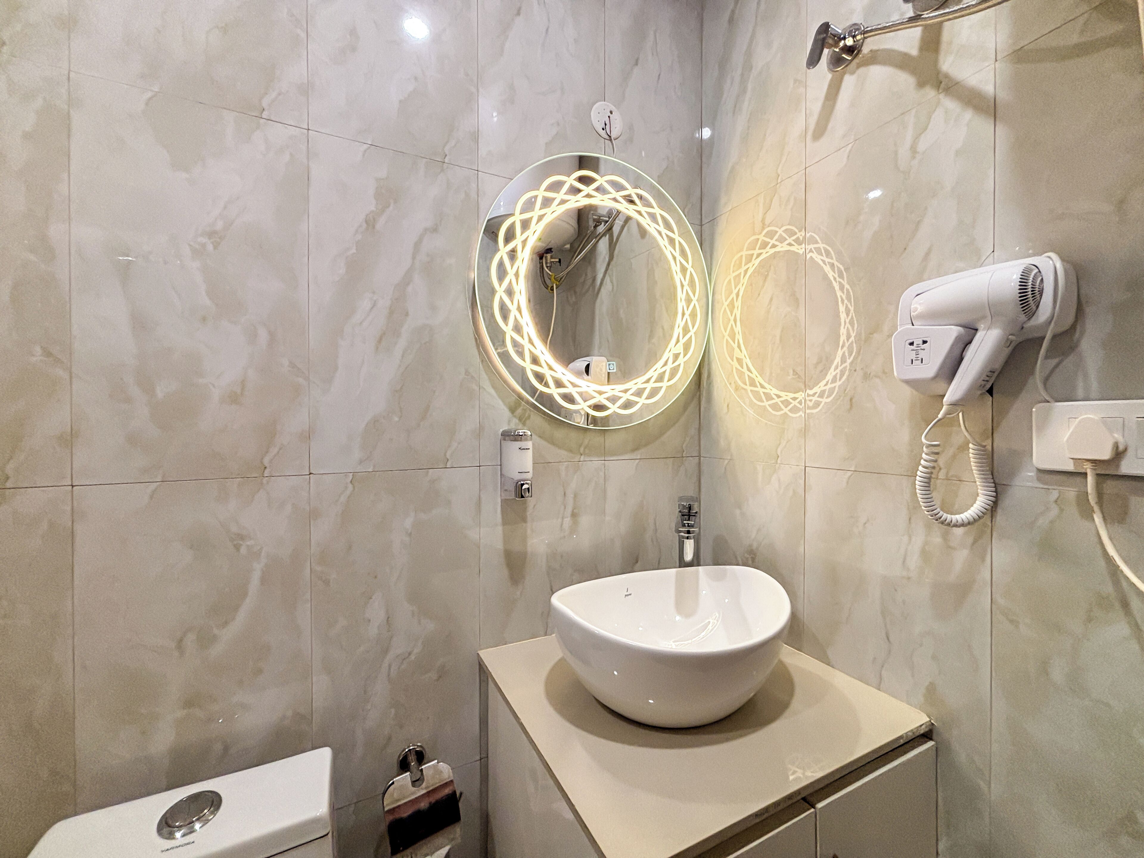 A bathroom with a white sink, a mirror with a circular light design, a hairdryer, and a towel dispenser.