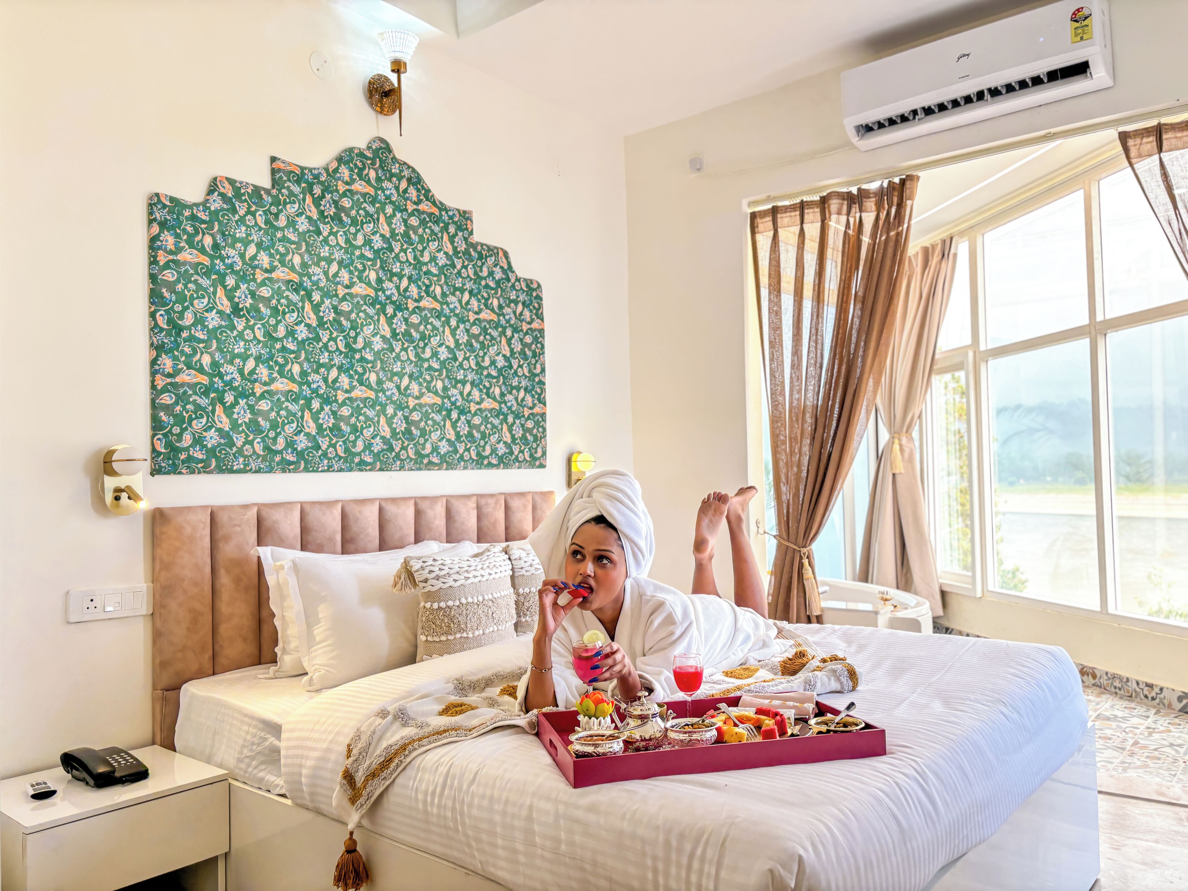 A hotel room with a bed, a bedside table with a phone, a decorative wall hanging, and a window with curtains.