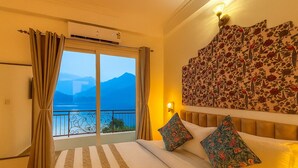 Hotelkamer met een grote schuifglazen deur die uitkomt op een balkon met uitzicht op de bergen. De kamer heeft een bed met kussens met bloemenmotief en een wandlamp.