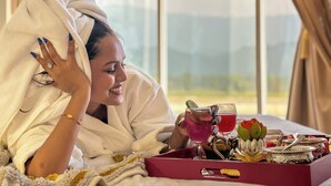 Iemand in een witte badjas, met een handdoek om het hoofd, ligt op een bed met een dienblad eten en drinken.