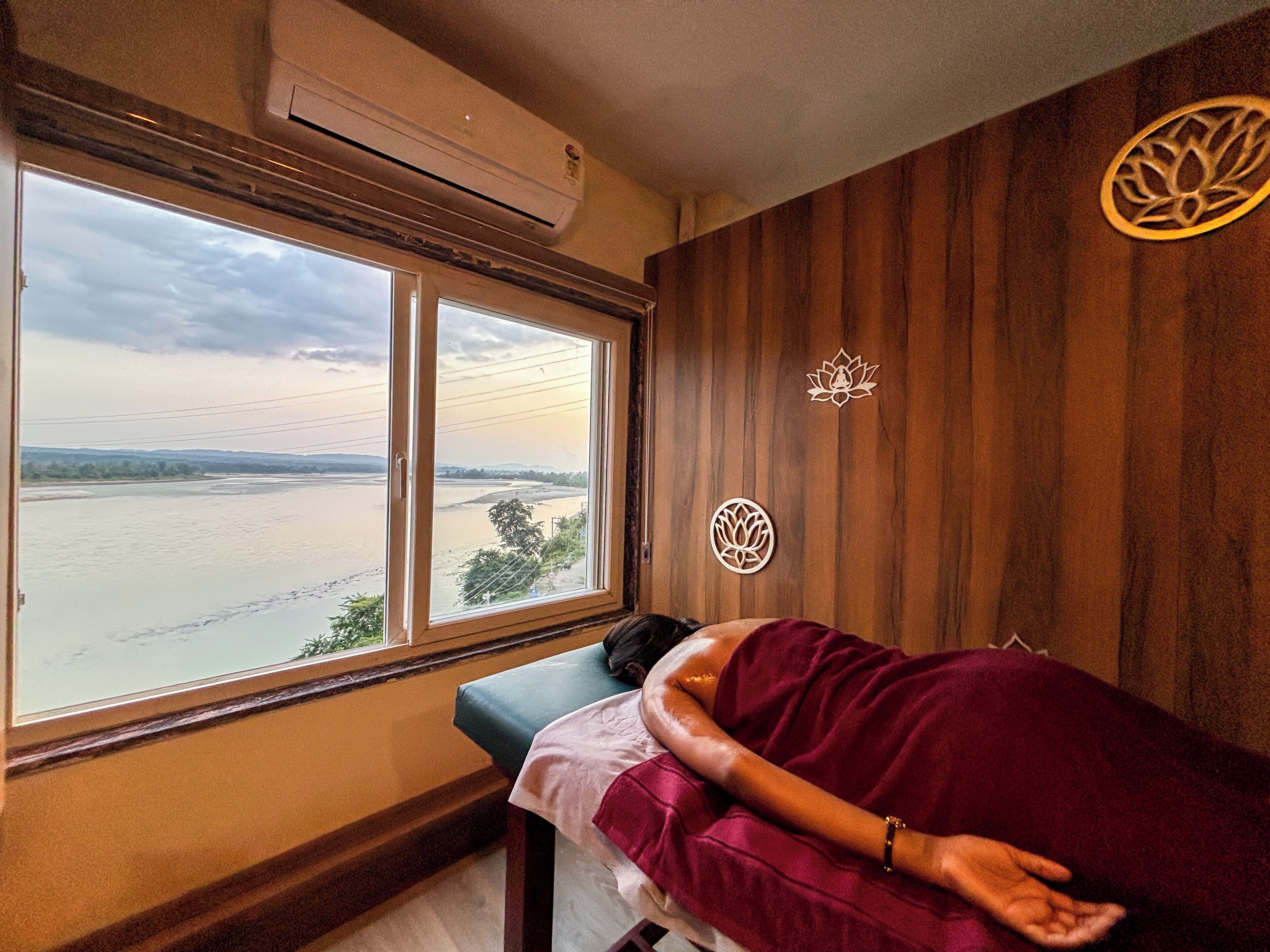 A room with a large window overlooking a river, a wooden wall with decorative elements, and a massage table with a person lying on it.