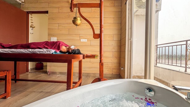 A spa room with a wooden massage table, a wooden massage chair, and a bathtub filled with water and rose petals. - The Neeraj Ganga Rajmahal Wellness Dream (Rishikesh)