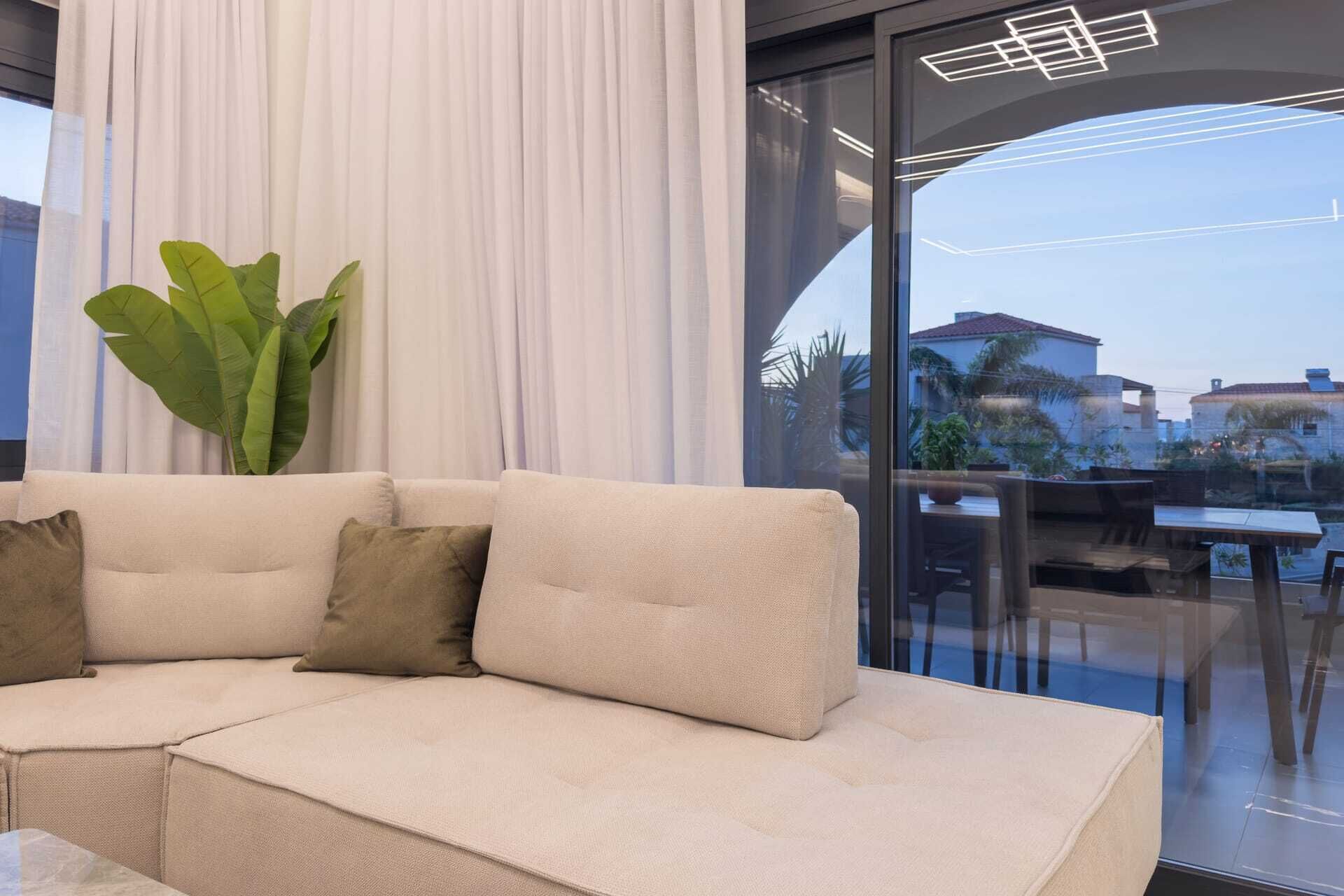 Ein modernes Wohnzimmer mit einem beigen Sofa, einer großen Grünpflanze und Blick auf eine Stadtlandschaft durch bodengrosse Fenster.