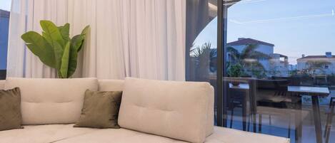 Ein modernes Wohnzimmer mit einem beigen Sofa, einer großen Grünpflanze und Blick auf eine Stadtlandschaft durch bodengrosse Fenster.