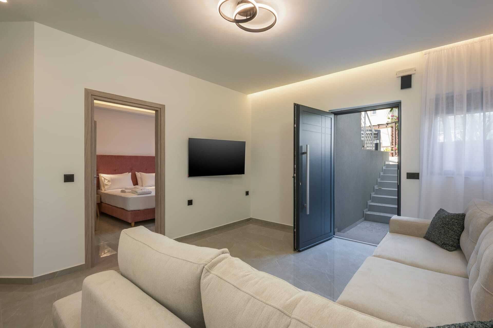 Ein modernes Wohnzimmer mit einer Sofagarnitur, einem Wand-Fernseher und einer Tür zum Schlafzimmer.