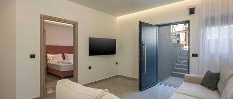 Ein modernes Wohnzimmer mit einer Sofagarnitur, einem Wand-Fernseher und einer Tür zum Schlafzimmer.