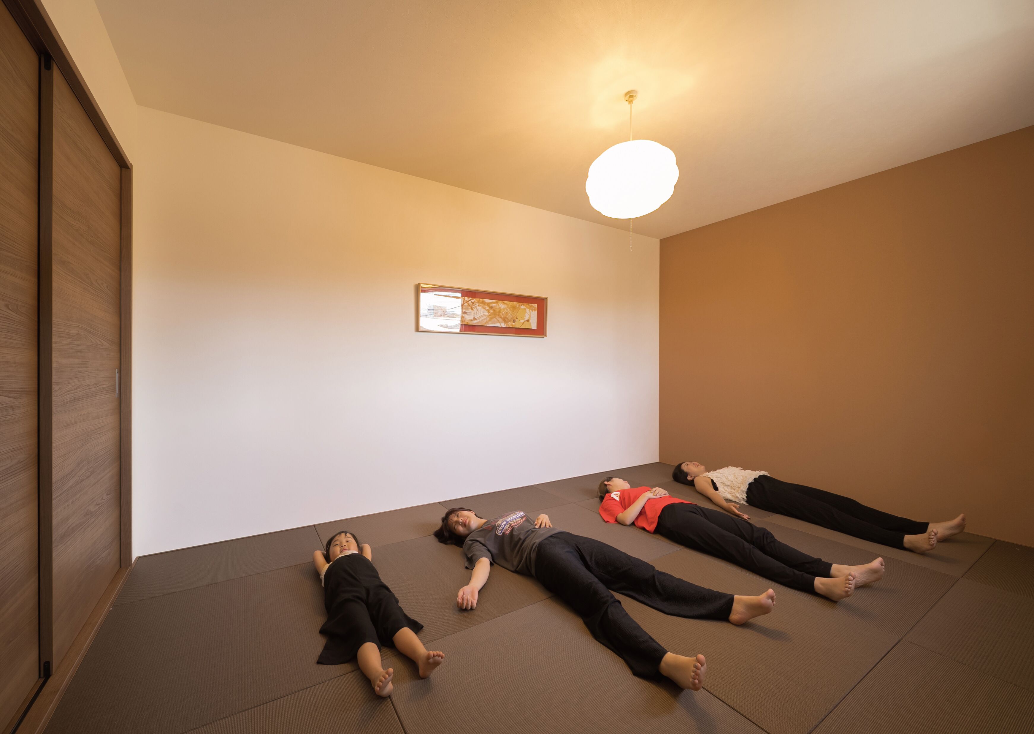 A room with four people lying on mats on the floor, a ceiling light, and a framed picture on the wall.
