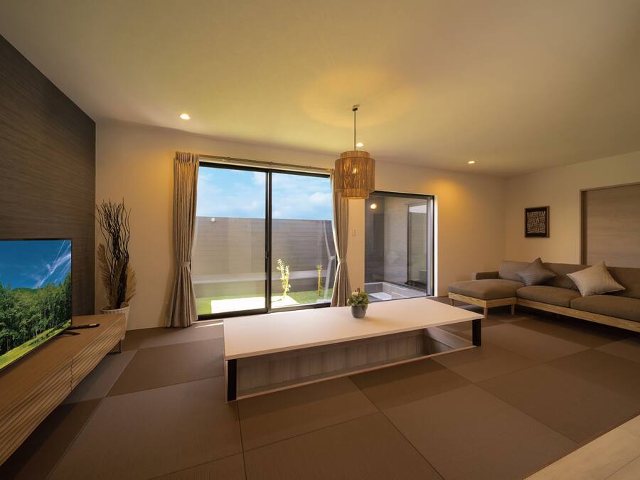 A modern living room with a large flat-screen TV, a sofa, a coffee table, and a view of a garden through large sliding doors.