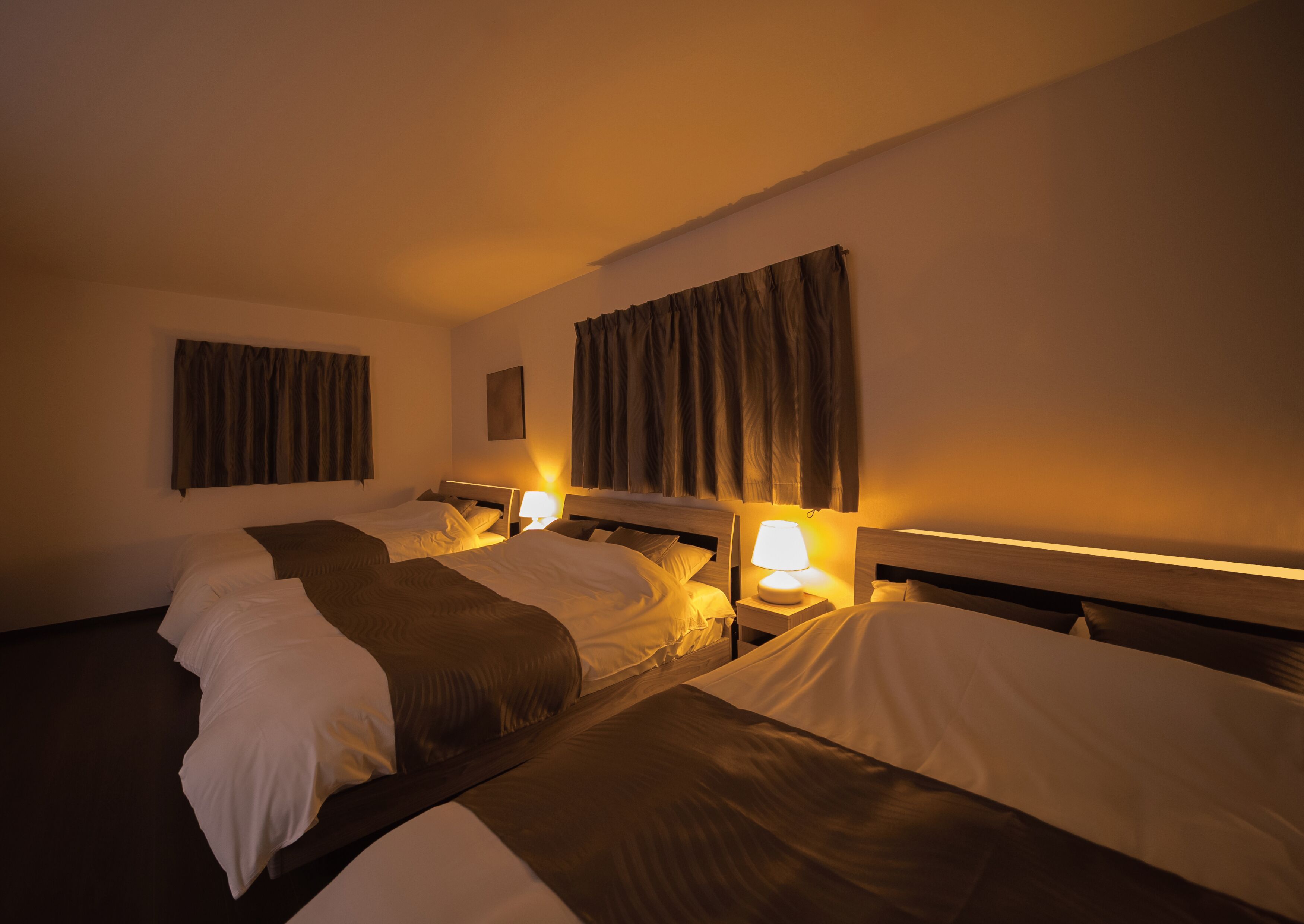 A hotel room with three single beds, each with a bedside table and lamp. The room has a simple, clean design with curtains on the windows and a small painting on the wall.