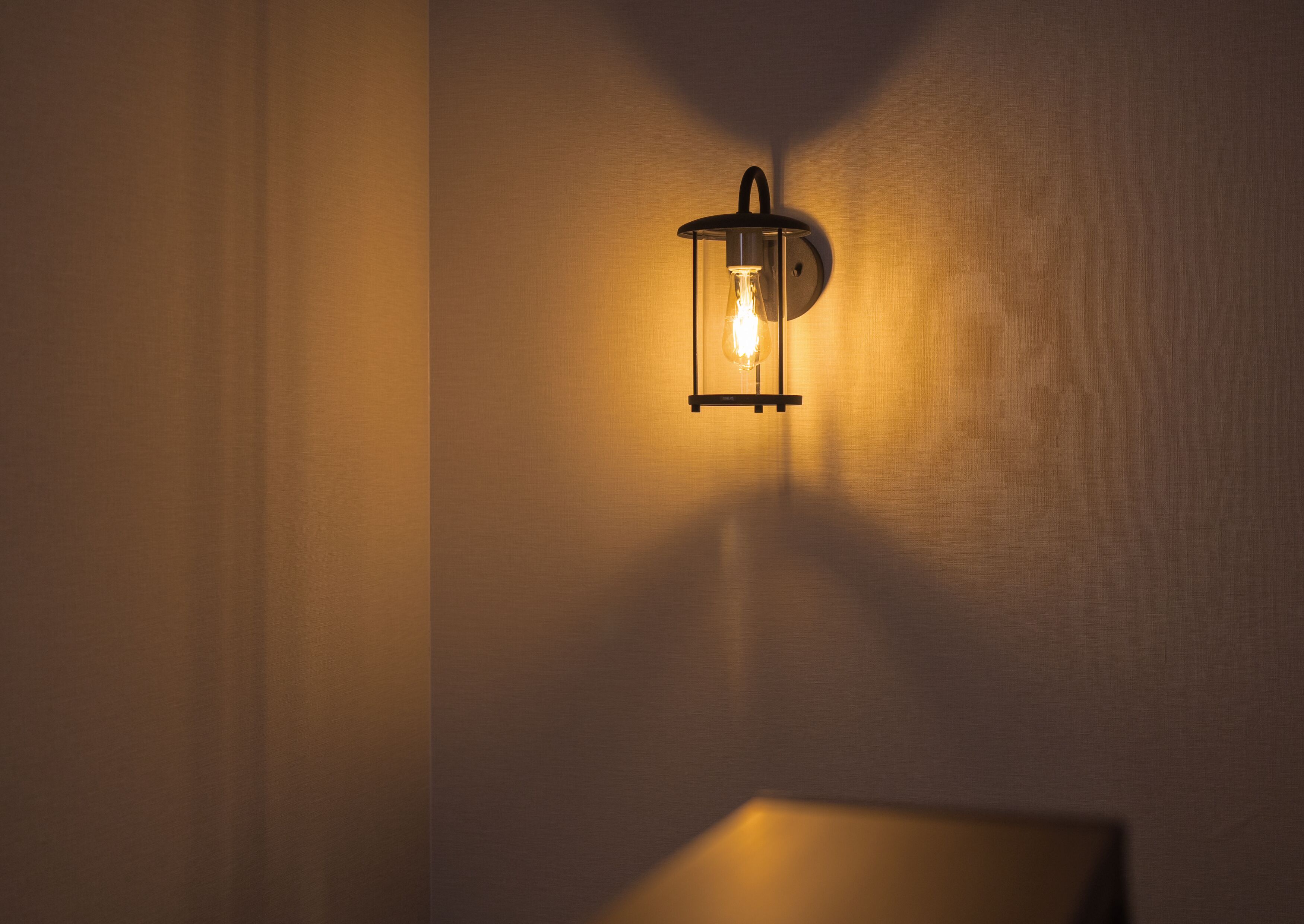 Una lámpara de pared con bombilla visible, proyectando un resplandor cálido y sombras sobre una pared con textura.